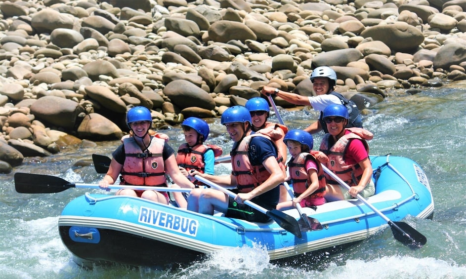 Een groep mensen in een blauwe Riverbug-mats peddelt over een rivier met een rotsachtige bodem. 3
