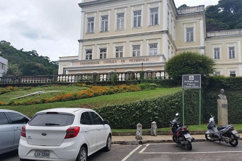 Ein weißes Auto vor einem Gebäude mit der Aufschrift 'Universidade Catolica de Petrópolis', einer Statue und einem Schild für 'Rei da Floresta'. 2