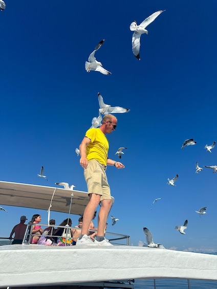 A man in a yellow shirt and beige shorts standing on a boat deck with seagulls flying around him. 5