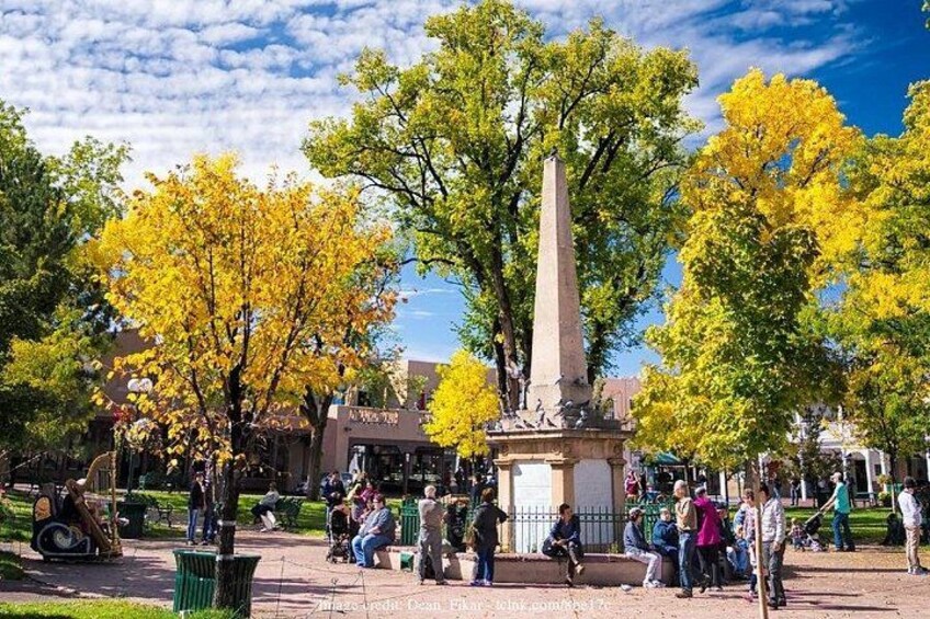 A park with a tall monument, trees with yellow leaves, and people walking and sitting. 5