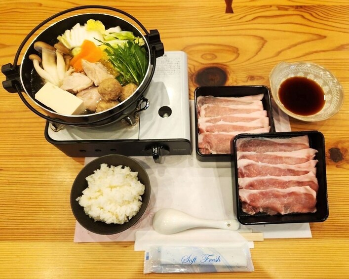 A meal setup with a pot of soup, rice, sliced meat, and condiments on a wooden table. 3