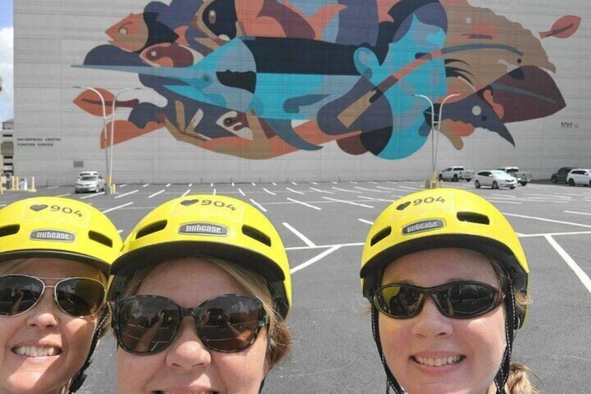 Three people wearing yellow helmets with the number 904, standing in a parking lot with a colorful mural in the background. 1