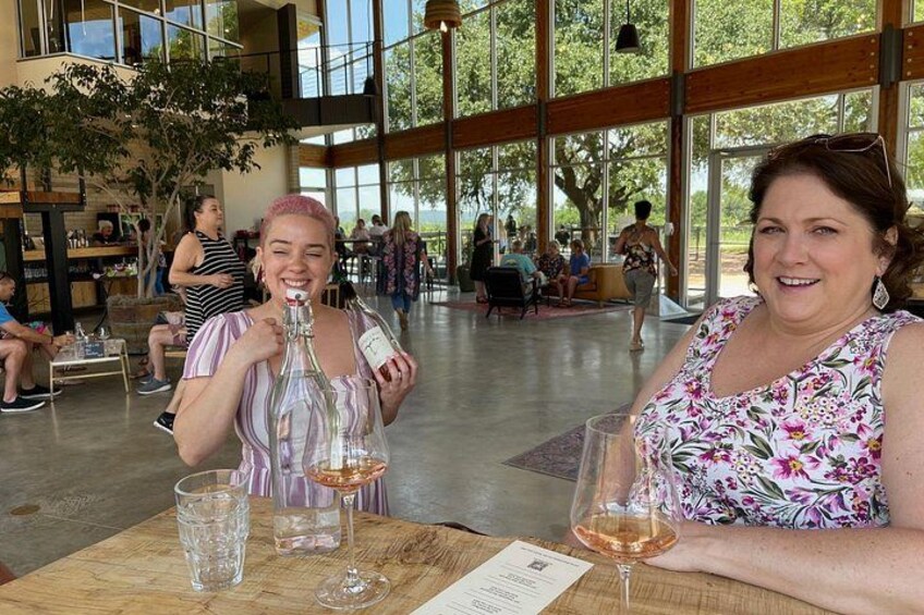 Two people at a table with wine glasses and a bottle, in a spacious indoor area with large windows and people in the background. 2