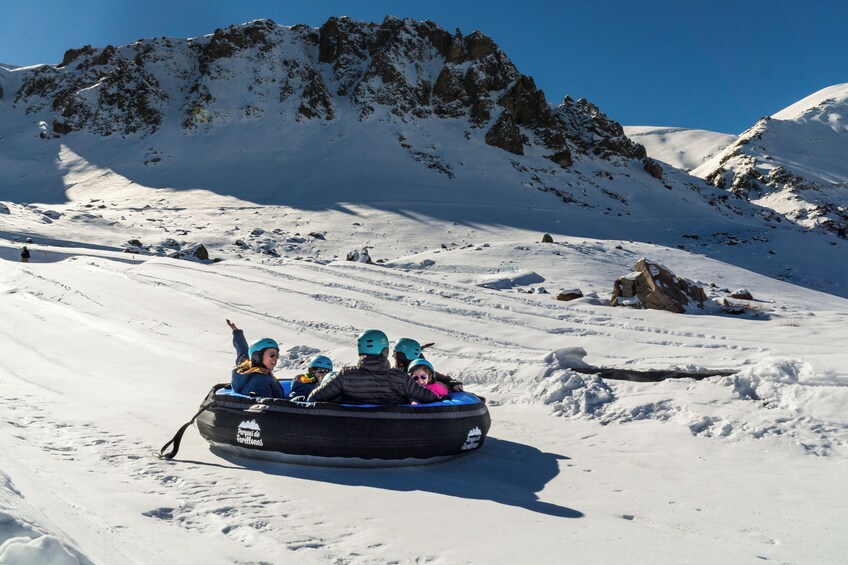 A group of people in a snow tube on a snowy mountain slope with rocky cliffs in the background. 3