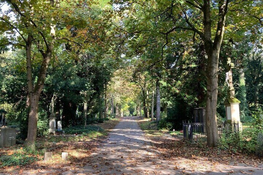 A tree-lined pathway in a park with tombstones and statues. 3