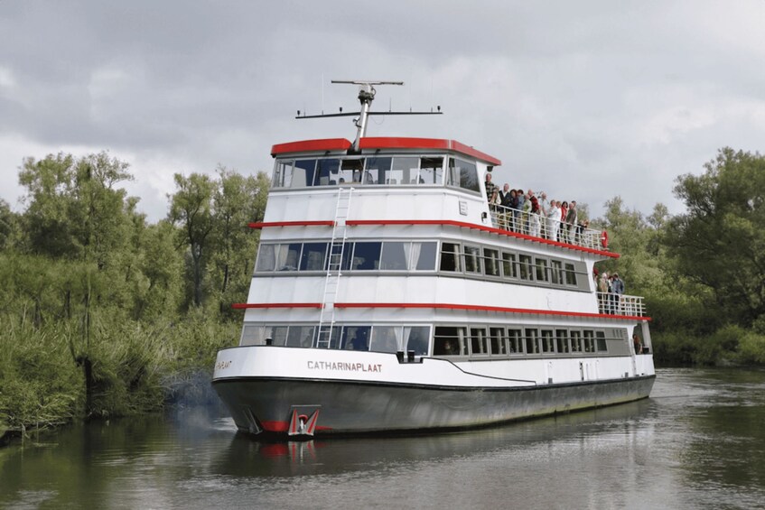 A white and red riverboat named 'Catharinaplaat' with passengers on deck, surrounded by greenery. 1
