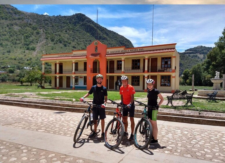 Three cyclists in front of a building with a red roof and columns, surrounded by greenery and hills. 1