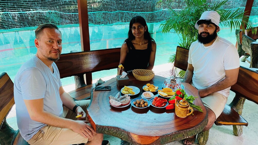Three people seated at a wooden table with a tropical setting, including a woven basket, plates of food, and a mug. 3