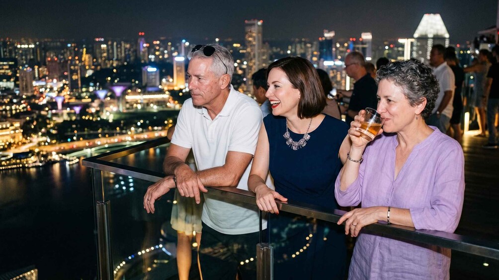 Three people standing on a balcony overlooking a cityscape with illuminated buildings and a river. 4