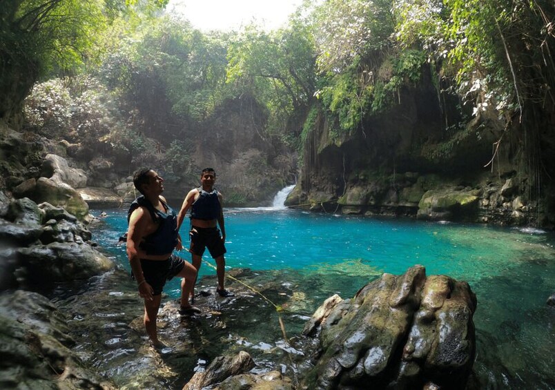 Two people standing by a clear blue pool surrounded by rocks and greenery. 2