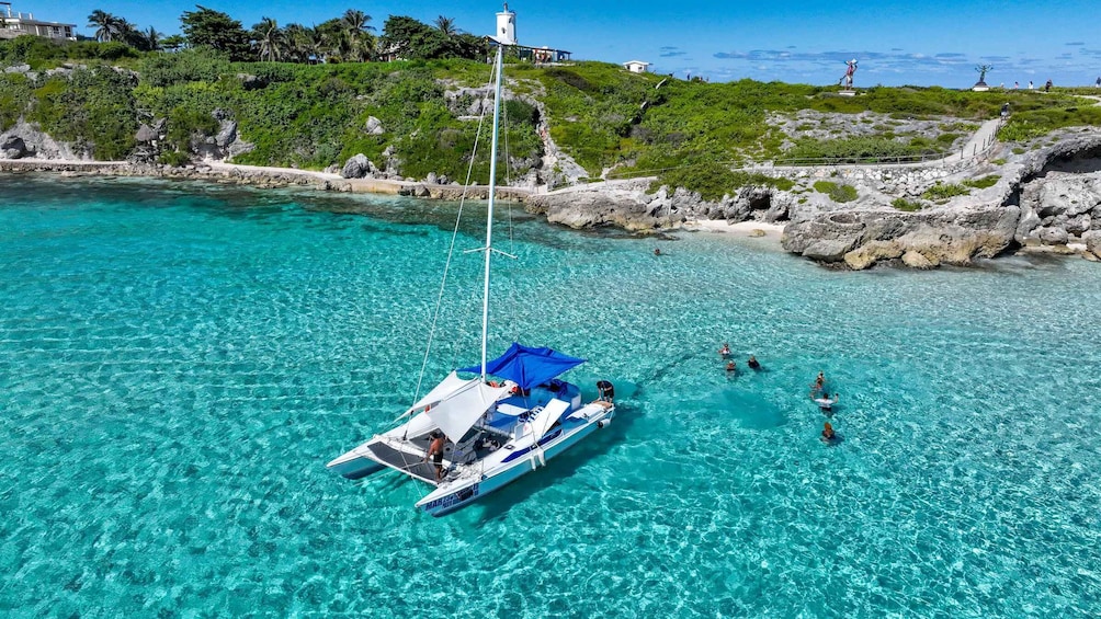 A sailboat anchored in clear turquoise waters with a rocky shoreline and lush greenery in the background. 1