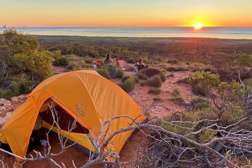 A desert landscape with a tent, rocky terrain, sparse vegetation, and a sunset. 1