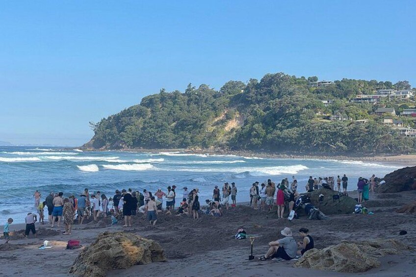 A crowded beach with people standing and sitting on the sand, waves crashing onto the shore, and a hill with buildings in the background. 4