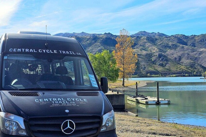 A black Mercedes van with 'Central Cycle Trail Co.' on the front, parked by a lake with mountains in the background. 2