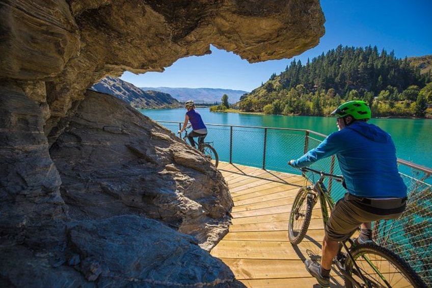 Two people on a wooden bridge over a lake, with a rocky cliff on one side and a forested hillside on the other. 3