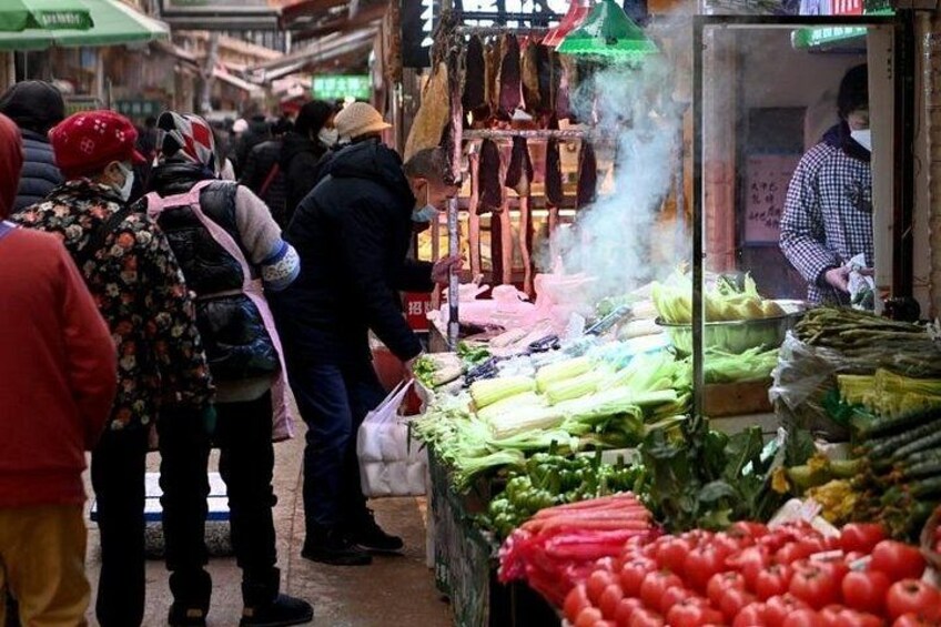 A bustling market with people shopping, various vegetables on display, and a vendor preparing food. 2