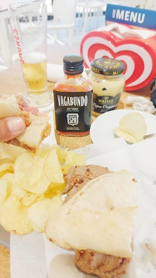 A sandwich with chips, a bottle of Vagarundo hot sauce, a jar of Maille Dijon mustard, and a glass of beer. 5