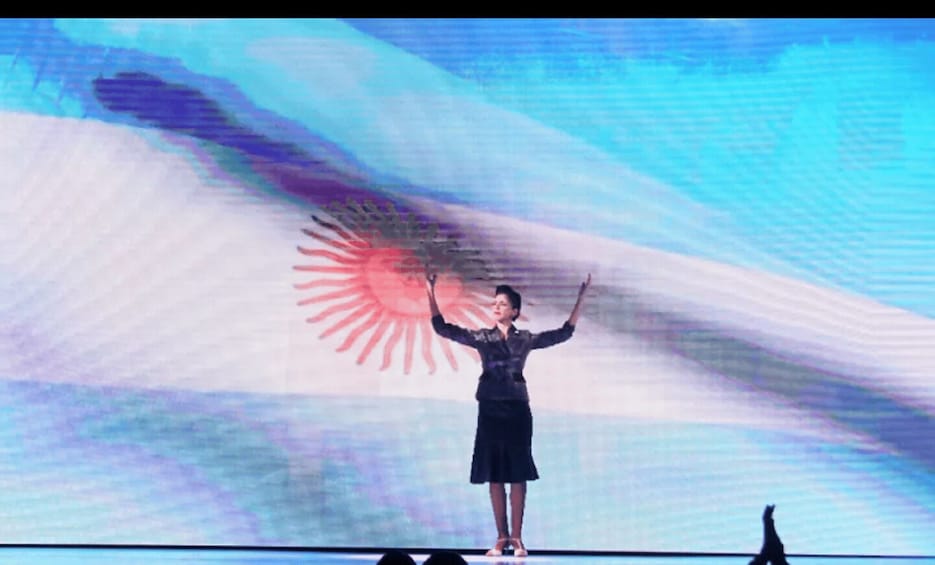 A person standing on a stage with a large screen displaying a flag in the background. 5