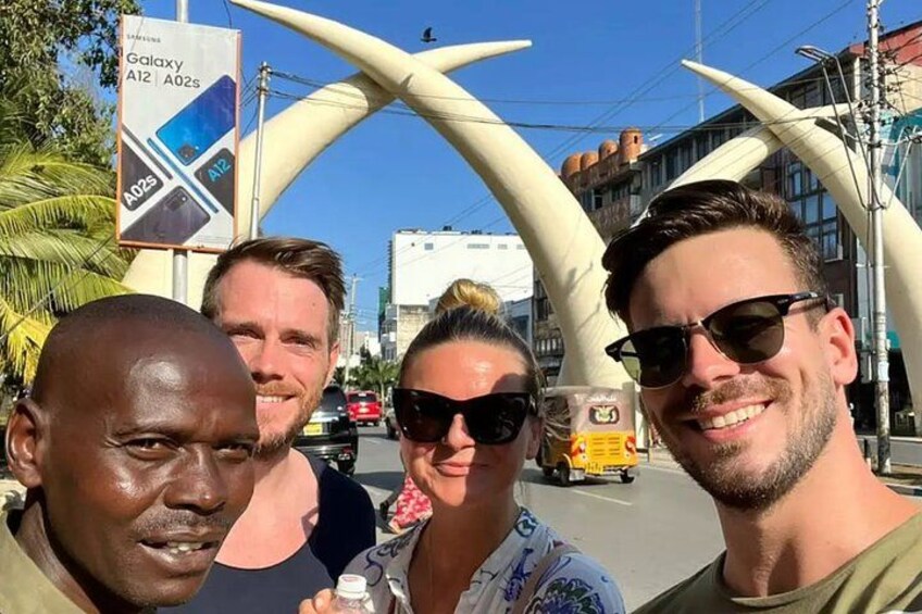 Un grupo de personas frente a una gran estructura en arco, con una calle y vehículos al fondo. 2