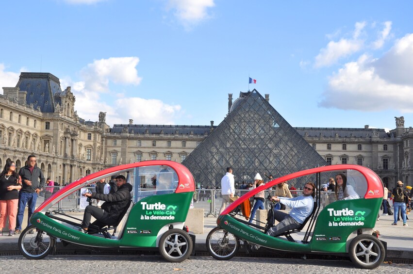 Two green and red 'Turtle Le Vélo-Cab' vehicles parked in front of the Louvre Museum with tourists around. 2