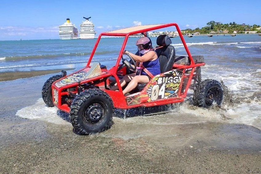 Een rode terreinwagen op een strand, met de oceaan en cruiseschepen op de achtergrond. 1