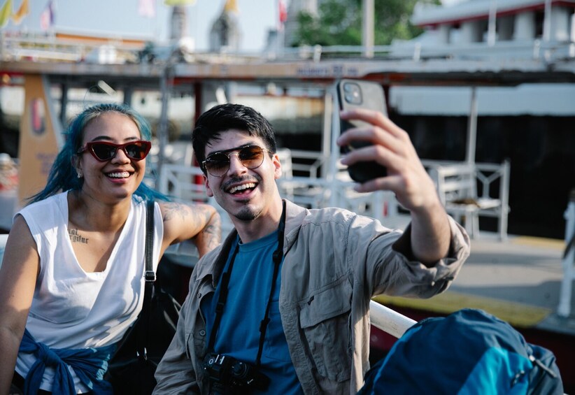 Twee personen maken een selfie met een smartphone, één met een tatoeage op hun arm, en een blauwe rugzak. 3