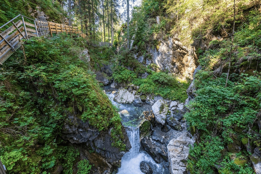 A forested gorge with a waterfall, rocky terrain, and a wooden bridge. 1