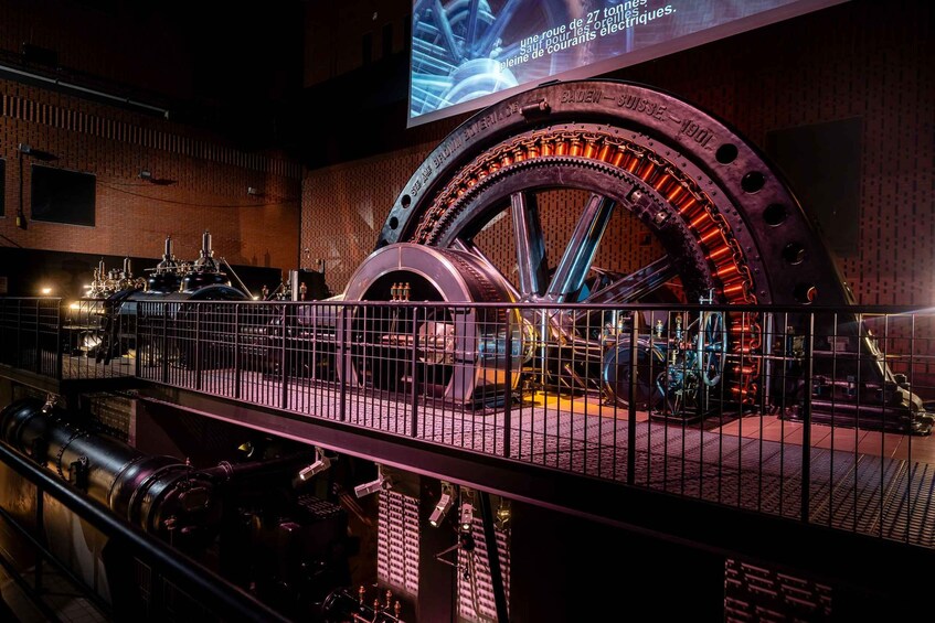A large mechanical wheel with intricate gears and chains, surrounded by industrial equipment and a brick wall. 1