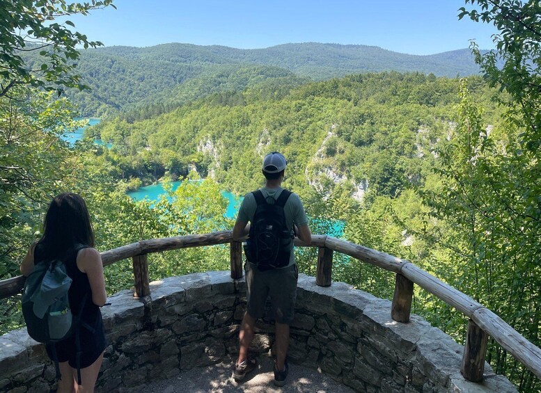 Dos personas en una plataforma de piedra con vista a un lago turquesa rodeado de colinas boscosas. 3