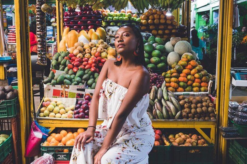 A person sitting in front of a fruit stand with various fruits and vegetables displayed. 1