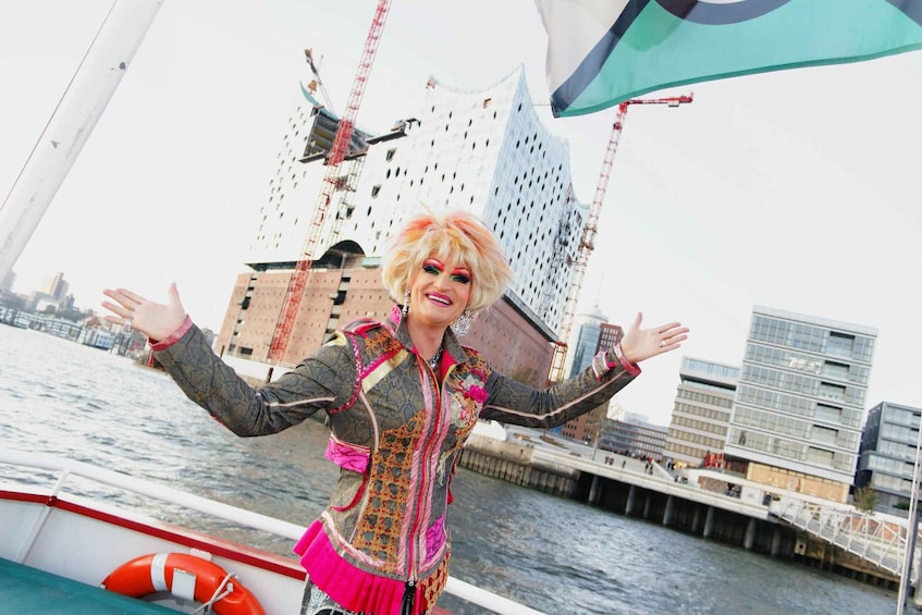 Een persoon in een boot met uitgestrekte armen, gekleed in een kleurrijke jas, met een stadsgezicht en gebouwen op de achtergrond. 1