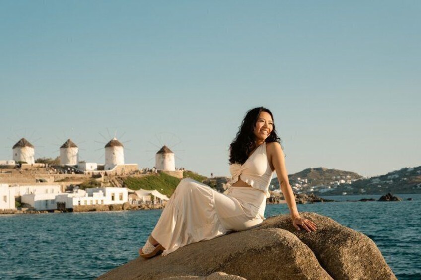 Una mujer con un vestido blanco sentada sobre una roca junto al mar, con molinos de viento al fondo. 1