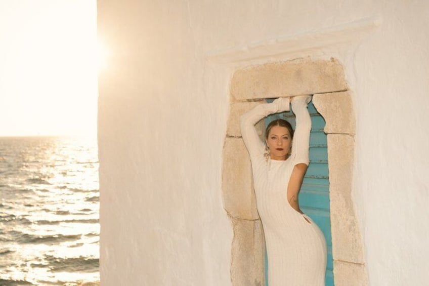 Una persona con vestido blanco apoyada en una pared de piedra con una puerta azul, cerca del mar. 4