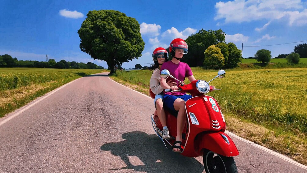 Two people on a red Vespa scooter on a rural road with fields and trees. 1