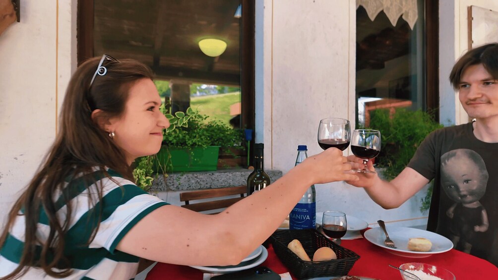 Two people toasting with wine glasses at an outdoor table with a basket of bread, a bottle of Manhva, and a plate with a small dish. 5