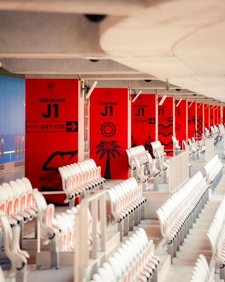 Filas de sillas de plástico blanco bajo un área techada con paredes rojas que muestran 'SEGURANE J1' y el logotipo de una palmera. 3