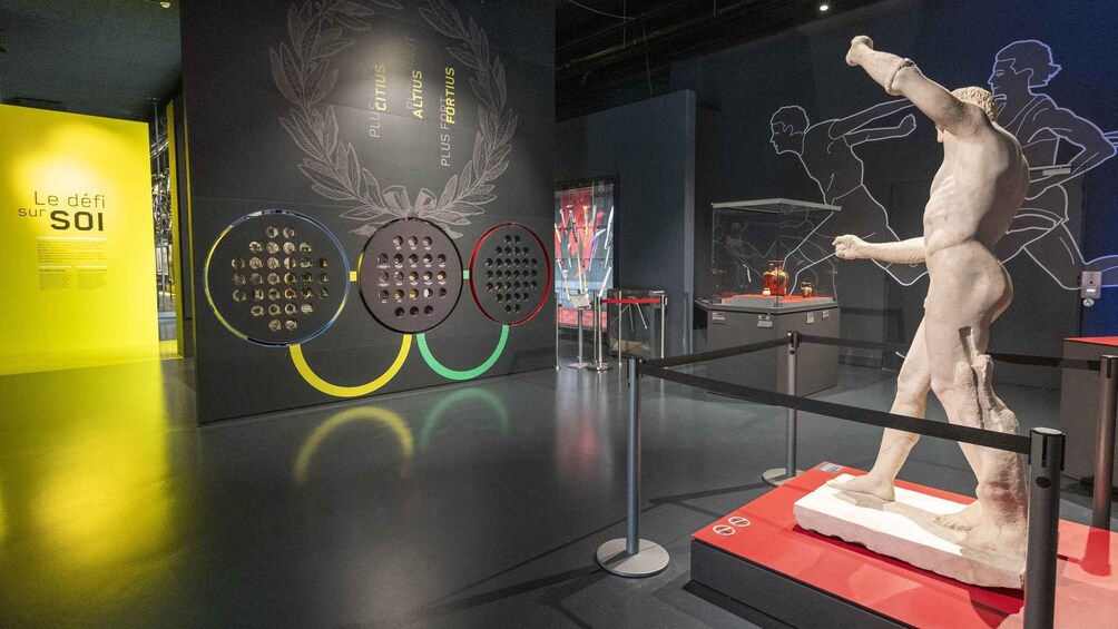 Una exhibición de museo con una escultura de figura masculina, anillos olímpicos y una pared con texto y gráficos. 4
