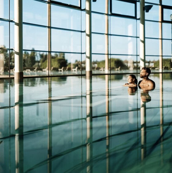 A modern indoor pool with glass walls reflecting the sky and surrounding trees. 2