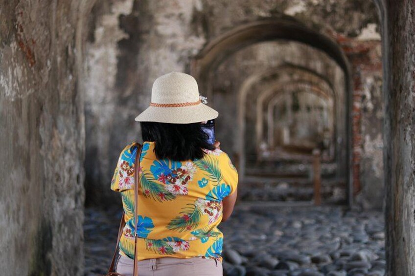 Una persona con una camisa floral amarilla y un sombrero de paja caminando por un antiguo corredor de piedra con arcos. 3