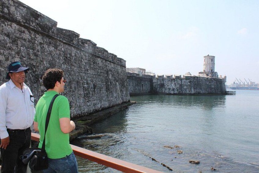 Un muro di pietra accanto a un’acqua calma, con una torre che si intravede in lontananza. 2
