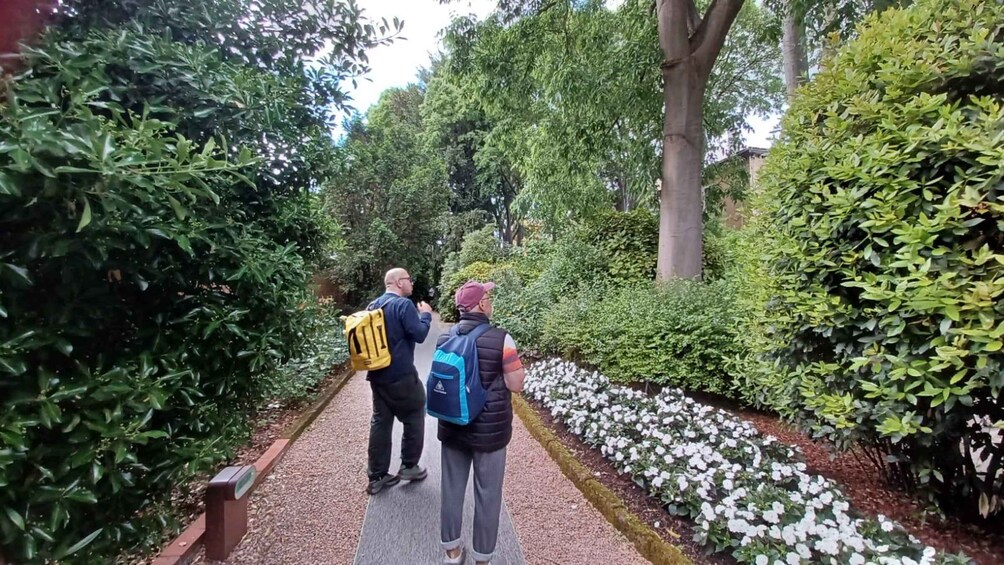 Two people walking on a path surrounded by dense greenery and white flowers. 5