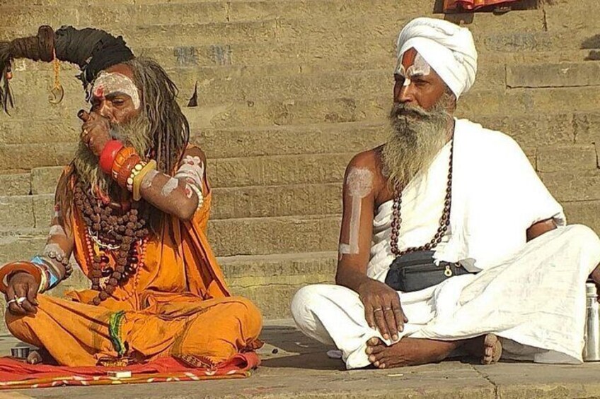 Two men sitting on the ground, one wearing orange and the other white, with a stone wall in the background. 4