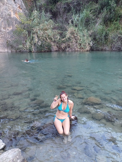 A woman in a blue bikini standing in shallow water with rocks, surrounded by greenery and a rocky cliff. 2