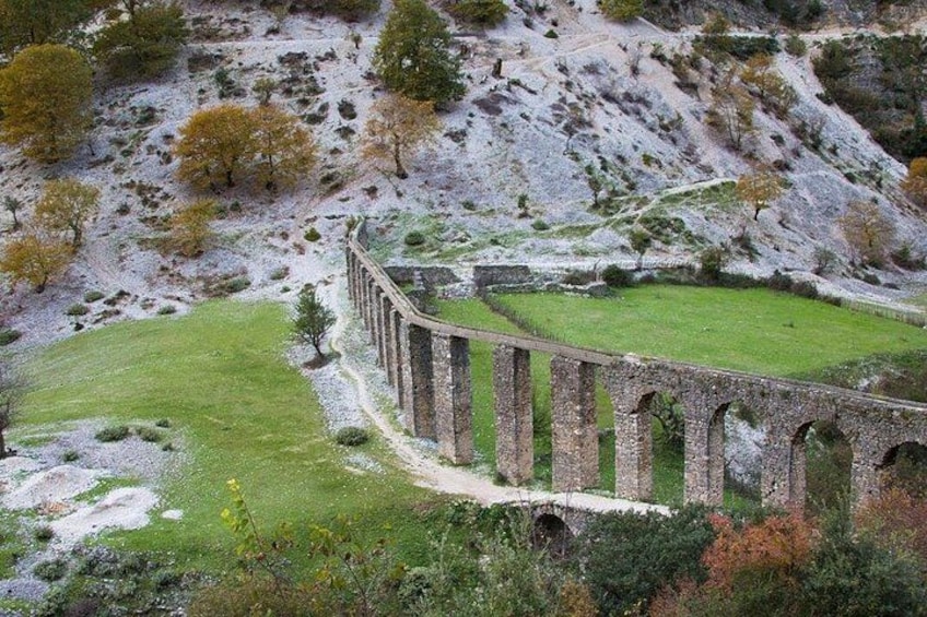An ancient aqueduct with arches, a grassy field, and a rocky hillside. 2
