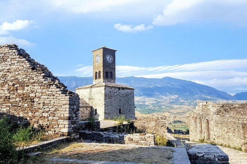 A historical stone building with a clock tower, surrounded by ruins and mountains in the background. 2