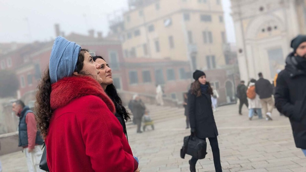 A person in a red coat and blue beanie walking on a cobblestone street with buildings in the background. 1