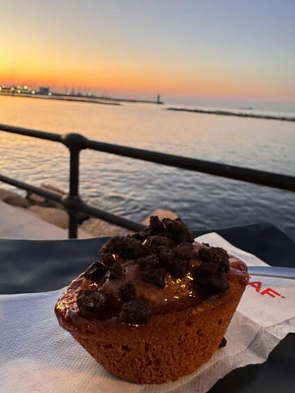 En muffin med chokladbitar på en servett, med en solnedgång och en brygga i bakgrunden. 3