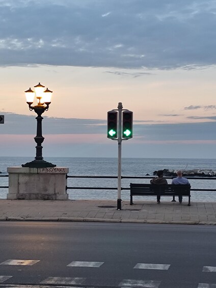 A street with a traffic light, a bench, a lamppost, and a body of water in the background. 2