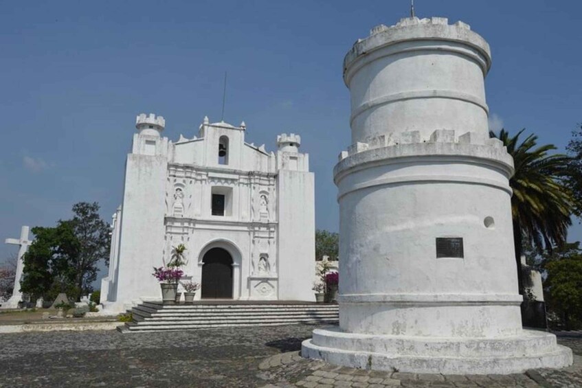 A white colonial-style church with a bell tower and a cylindrical monument in front. 1