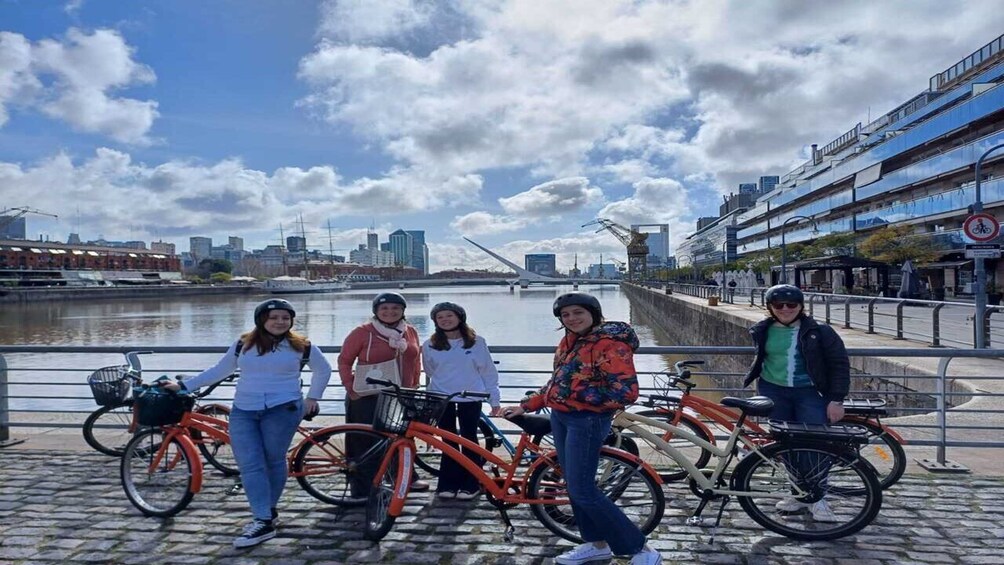 Un grupo de personas junto a bicicletas cerca de un muelle, con edificios y un puente al fondo. 1